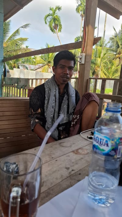 The image depicts a man sitting at a wooden table, likely outdoors. He is wearing a dark shirt with intricate patterns and a light-colored scarf around his neck. His right leg is crossed over his left. In front of him, on the table, is a glass with a straw, and a partially filled bottle of Aqua water. The background shows a structure with wooden planks and a corrugated metal roof. Beyond that, palm trees and a cloudy sky are visible, suggesting a tropical environment. The man appears to be looking towards the camera with a slight smile. Based on the context of the Grong-grong city, Indonesia, this scene likely captures a moment of relaxation or a casual meeting in a local establishment or a residence in a tropical area.