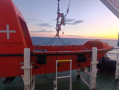 An orange lifeboat is suspended by a crane over the deck of a ship at dusk. The lifeboat is attached to the crane by multiple cables and rigging. The ship's deck is green and has white railings. The ocean is visible in the background, with a colorful sky. The scene suggests a maritime operation or maintenance activity on a vessel at sea, likely in or near Larsmo, Finland, given the context.