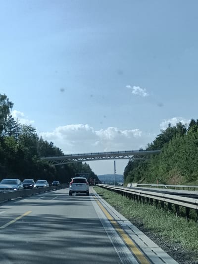 The image depicts a view from a car traveling on a highway in Hrusice, Czechia. The road is paved with asphalt, marked with yellow and white lines, and bordered by a metal guardrail and grassy embankments on the right. Trees and lush green foliage line both sides of the highway, creating a scenic, natural setting. In the distance, an elevated bridge, possibly part of the highway infrastructure, spans across the view. Several cars are visible on the road, suggesting ongoing traffic. The sky is a clear, light blue with a few scattered white clouds, indicating fair weather. The sunlight casts shadows on the road, implying it is daytime, likely late morning or early afternoon. A white SUV is prominently featured in the foreground, with its license plate partially visible. The overall impression is one of a pleasant drive through a green, hilly landscape.