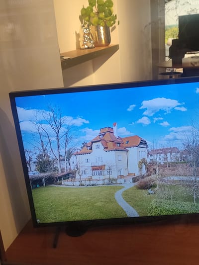 The image displays a television screen showing an aerial view of a large, traditional European-style house with a red-tiled roof. The house is surrounded by a well-maintained green lawn, bare trees, and other smaller buildings in the background. The sky is bright blue with fluffy white clouds. A winding stone pathway leads from the front of the house through the lawn. The scene appears to be captured in late autumn or early spring, given the lack of foliage on the trees. The television itself is placed on a wooden surface, likely a table or desk. The background behind the TV shows a blurry reflection, possibly of a window or another room, with a shelf holding a decorative plant and a metallic bunny figurine. The television screen is reflecting light from the surrounding environment, creating glare and obscuring some details.