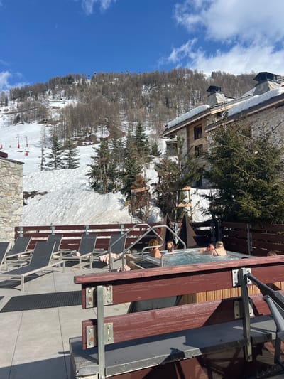 The scene is a rooftop patio in Val-d'Isère, France, at the coordinates 45.442, 6.977. There are four people relaxing in a hot tub, a young girl, and two women, and a man, all wearing swimwear. The hot tub is positioned in the center of the patio, surrounded by wooden fencing, and a wooden railing runs around the edge. The hot tub is filled with steaming water. A couple of lounge chairs, some with cushions, are laid out on the patio. Beyond the patio, the snow-covered slopes of a ski resort stretch upwards to meet a line of trees, a blue sky dotted with clouds, and a ski lift in the background. The buildings in the scene are built in a traditional stone and timber style. The patio floor is concrete, with a grey mat placed in front of the hot tub. The patio is located on a roof with a view overlooking the slopes.