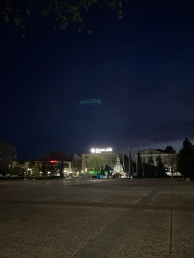 The image captures a nighttime scene in Nova Zagora, Bulgaria. A large, paved public square dominates the foreground, stretching towards illuminated buildings in the middle ground. A sign for "BANKA DSK" is visible in bright lights. Several other buildings are lit up, suggesting a commercial area. The sky is dark blue. The upper right edge shows a building with lit windows and green foliage near it. Along the left edge are more tree branches against the night sky. A faint, long, streak-like object appears in the sky.