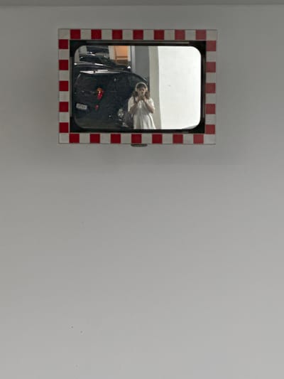 In Höhenkirchen-Siegertsbrunn, Germany, a security mirror with a red and white checkered frame is mounted on a wall. The mirror reflects a person taking a selfie with a phone. Behind the person, dark-colored cars are visible, suggesting the mirror is in a parking area, likely a garage. The background of the mirror's reflection is illuminated by artificial light, with a white wall and a portion of a structure visible. The overall scene is indoors, under artificial lighting.