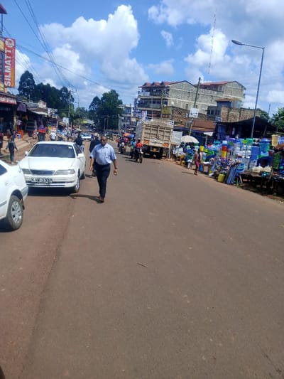 The image depicts a daytime street scene in Kangema, Kenya, under a blue sky with scattered white cumulus clouds. A two-lane paved road, appearing dark grey-brown, extends from the foreground into the distance.

In the foreground, a male adult wearing a light blue collared shirt, dark trousers, and dark shoes walks along the road, moving from left to right. To his left, a white sedan with license plate KAW 333P is parked on the unpaved reddish-brown shoulder, partially obscuring another white vehicle.

Along the left side of the road, commercial buildings are visible, one featuring a prominent vertical red and yellow sign labeled "SILMATT." Numerous market stalls with awnings display merchandise, and several pedestrians are present. Overhead utility lines crisscross above the street.

On the right side of the road, market stalls prominently display a variety of colorful plastic household goods, including buckets, basins, and stools. Further down the road, a light-colored box truck is visible, along with multiple motorcycles and other vehicles. Multi-story buildings, constructed from concrete and stone, line the background on both sides of the street. A tall street light stands on the far right. Green trees are visible in the background, behind the buildings and in the distance.