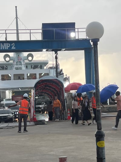 The image depicts a group of people disembarking from a ferry in Don Sak, Thailand. The time of day appears to be late afternoon or early evening, as indicated by the soft, diffused light. The sky is overcast, suggesting possible rain, which is further evidenced by the presence of several umbrellas, including blue and red ones.
The primary subjects are the passengers and ferry crew. Many passengers are seen holding umbrellas, some wearing life vests. A few individuals in the foreground are wearing high-visibility vests, likely indicating they are part of the ferry crew or port staff, guiding the disembarkation. A large ferry dominates the background, with its ramp extended to the pier. The ferry has multiple decks and windows.
The setting is a ferry terminal or port. Visible objects include the ferry itself, the pier infrastructure, a lamppost with a white globe light, traffic cones, and cars waiting on the pier. The ground is a concrete surface.
The activity is the process of passengers and vehicles leaving the ferry. There is a sense of organized movement as people make their way off the vessel. The mood is somewhat subdued, possibly due to the overcast weather and the necessity of using umbrellas.
Text visible in the image includes "AMP 2" on the side of a blue structure near the ferry. There is also a yellow sticker on the lamppost with some text that is difficult to read clearly but appears to be in Thai script.