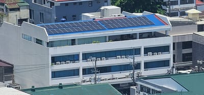 The image shows an elevated view of a multi-story white building with solar panels installed on its blue-roofed upper level. The building has multiple floors with large, blue-tinted windows. Several outdoor air conditioning units are visible on the exterior walls. A prominent sign with the word "actron" is visible on a lower level. In the background, other buildings and trees are present. The context suggests this is likely a commercial or office building. Based on the information available, this building is located in the central business district of Makati City, Philippines, a bustling urban environment characterized by numerous commercial structures.