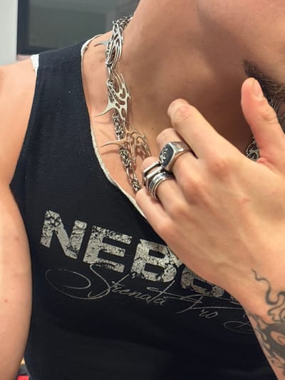 A close-up, angled photograph captures a male individual indoors, identified as being in Asnières-sur-Seine, France. The subject's left chest, neck, partial jawline with stubble, and right hand are visible. He wears a black tank top with a distressed white logo displaying "NEBBI" and "Strenata Pro". Around his neck is a silver-colored chain necklace composed of multiple ornate, tribal-style links with pointed elements. His right hand, positioned near his chin/jawline, features a large silver signet ring on the ring finger and two stacked silver rings on the pinky finger. A black tribal-style tattoo is visible on the inner right forearm/wrist, and a thin, wavy line tattoo is on the back of the right hand. The background is blurred, indicating an interior setting with indirect lighting.