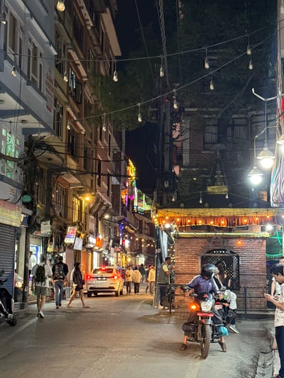 This is a night scene in Kathmandu Metropolitan City, Nepal. A street is lined with buildings of varying heights, many with multiple stories and windows. The buildings are densely packed together, with a mix of architectural styles. Power lines and street lights crisscross above the street, adding to the cluttered urban landscape.
People are walking along the street, and a motorcycle is parked on the side. A tuk-tuk or similar vehicle is in the upper right. The lighting is a mix of artificial street lights and the glow from shops and businesses, creating a warm but slightly chaotic atmosphere.
The street is paved, and there is evidence of pedestrian activity. The image captures the bustling atmosphere typical of a city in Nepal at night.