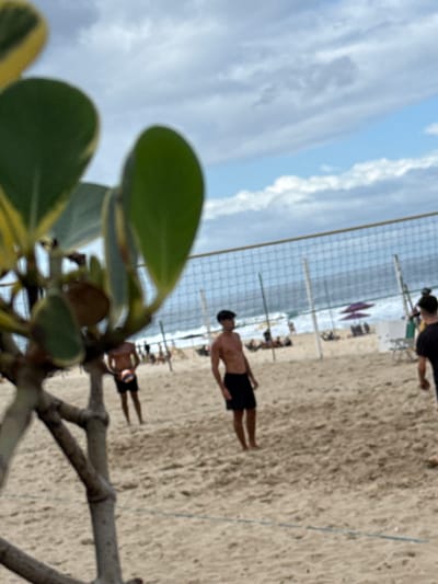 The image displays a beach scene in Rio de Janeiro, Brazil, captured under a partly cloudy sky. In the extreme foreground, a blurred plant with green and yellow variegated leaves and multiple branches partially obscures the left side of the frame. The midground features a sandy beach with a beach volleyball net spanning horizontally across the scene. To the left of the net, a shirtless male wearing black shorts stands, facing right, holding a multi-colored volleyball. Another shirtless male, also in dark shorts, is partially visible on the far right, facing left. Further in the background, several indistinct human figures are distributed across the sand, with two beach umbrellas (one purple, one white) also discernible. The background shows the ocean with visible white waves breaking near the shoreline, beneath a sky characterized by scattered white and grey clouds.