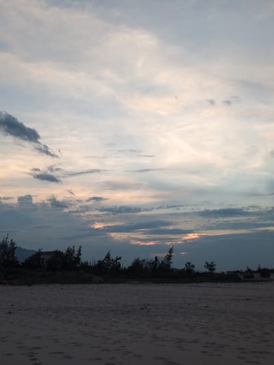 The image depicts a serene beach scene at dusk in Hoành Sơn Ward, Vietnam. The foreground is dominated by the textured sand of the beach, with subtle footprints and debris scattered across its surface. In the middle ground, a line of dark silhouettes of trees and vegetation runs along the horizon, interspersed with a few distant buildings, including a notable white structure with a red roof. The silhouette of a mountain is visible on the far left. The sky above is a dramatic canvas of clouds, with varying shades of gray, white, and hints of orange and pink, suggesting the fading light of sunset. The overall atmosphere is tranquil and natural, with no people or active interactions visible.