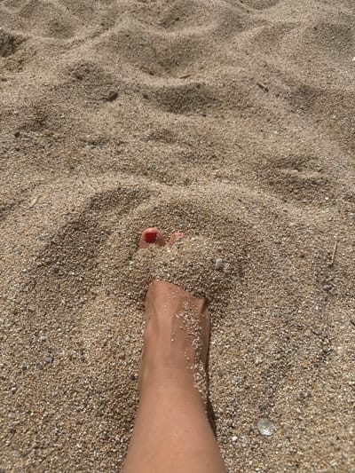 The image shows a person's feet buried in the sand. One foot is mostly visible from the ankle down, covered in sand grains. The toes of the other foot are peeking out of the sand and are painted with red nail polish. The environment is a sandy beach, consistent with the coastline of Badalona, Spain. The sand is a light brown color with visible pebbles and small stones. The lighting suggests it is daytime, likely sunny.