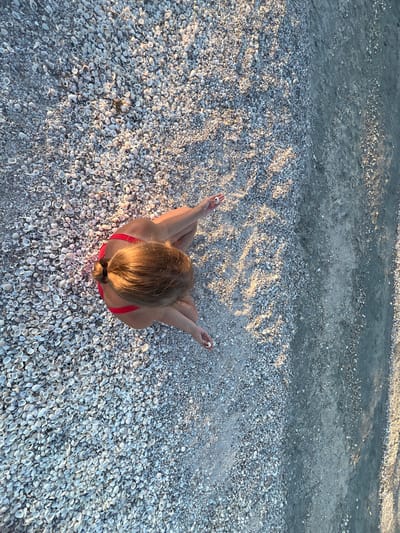 The image shows a top-down view of a person sitting on a beach covered in shells. The person is wearing a red bikini and is seated in a cross-legged position with their hands resting on their knees, palms up, in a meditative pose. Their hair is blonde and pulled back in a ponytail. The beach is densely covered with small, light-colored seashells, creating a textured surface. The water's edge is visible at the top of the frame, where the water is calm and appears to be shallow. The lighting suggests it is either early morning or late afternoon, as indicated by the long shadows cast on the sand and shells. The overall mood is peaceful and serene. The location is specified as Мысовское сельское поселение, Ukraine. There is no visible text in the image.