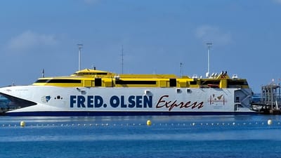 A Fred. Olsen Express ferry, featuring a white hull with a blue waterline stripe and a bright yellow superstructure, is observed stationary in the port of Los Cristianos, Spain. The vessel's hull prominently displays "FRED. OLSEN" in blue and "Express" in red script, along with a circular blue and white logo at the bow and an orange stylized island map near the stern. Its sleek design incorporates a pointed bow and dark-tinted windows across the yellow upper decks, from which multiple antennas and masts ascend. The stern is positioned directly alongside a grey metallic pier structure, implying a docked or moored state. The foreground comprises calm, deep blue water, segmented by a horizontal line of small yellow buoys and a single larger yellow buoy behind it. The background consists of a clear, light blue sky with negligible cloud presence. No individuals are discernible within the scene.