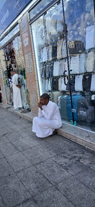 The image displays an outdoor street scene in Medina, Saudi Arabia, specifically a sidewalk adjacent to a commercial storefront. A partial sign in the upper left corner confirms the location with the visible text "...madinah" and Arabic script. In the foreground, a male with dark skin, dressed in a white thobe and dark shoes, is squatting on the dark gray tiled sidewalk, leaning against the base of a large glass display window. He is actively engaged in a phone call, holding a dark smartphone to his right ear with his right hand. His left hand rests on his left knee. To the left and further back, a second male, also wearing a white thobe, stands near the storefront, facing inward or slightly away from the camera. The storefront window displays various items, including numerous folded shirts and garments of different colors, as well as multiple suitcases in shades of blue, gray, and black. A black multi-hook stand is visible within the display. Reflections in the glass show a blue sky with white clouds and the upper portion of a distant building featuring square, arched windows.