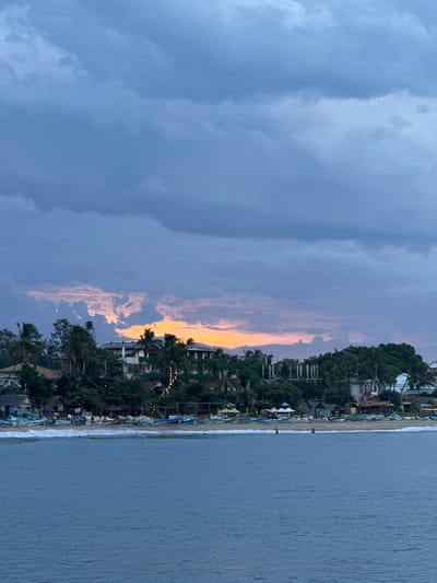 The image captures a scenic view of Arugam Bay, Sri Lanka, during what appears to be dusk. The scene is dominated by the ocean on the right and a dramatic sky on the left, with a coastal settlement nestled between them. The sky is a gradient of colors, transitioning from a deep, dusky blue on the left to lighter shades, with streaks of soft orange and pink where the last rays of sunlight are breaking through the clouds.
The shoreline is lined with numerous small, colorful boats pulled up onto the sand. Beyond the beach, a dense collection of buildings and lush green trees forms the coastal settlement. Some of these structures appear to be accommodations, with visible windows and balconies, while others might be shops or restaurants. String lights are visible, suggesting the area is preparing for the evening. The overall atmosphere conveyed is one of tranquility and the peaceful winding down of a day, bathed in the soft, diffused light of sunset. The gentle waves lapping at the shore and the distant lights create a serene ambiance. There are a few scattered people visible on the beach and near the water's edge. The weather appears calm, with the clouds adding a beautiful texture to the sky.