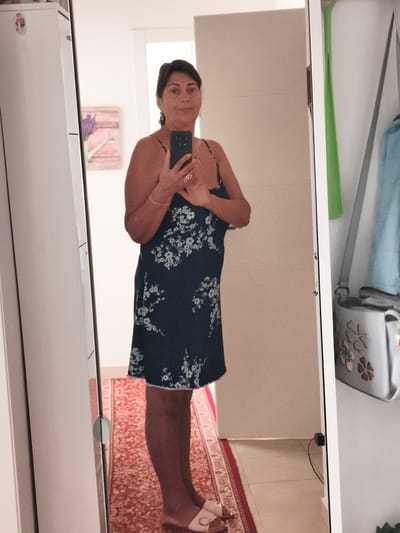 A full-body, front-facing selfie is taken by a woman in what appears to be a home environment, possibly an apartment in Livorno, Italy. The woman stands in front of a large, full-length mirror, reflecting her image. She is wearing a dark blue, sleeveless dress with a delicate white floral pattern. She is holding a smartphone in her right hand, angled to capture her reflection, and her left hand is resting on her chest, adorned with rings. The woman has medium-length, dark hair styled loosely around her shoulders and appears to be wearing light-colored slip-on shoes.

The background visible in the mirror includes a portion of a tall, white cabinet or closet on the left, a framed piece of art with lavender on the wall behind her, and a textured, light-colored wall on the right, possibly a door or another cabinet. A red patterned rug is visible on the floor beneath her. To the right of the woman's reflection, the mirror also shows a glimpse of a grey handbag with floral embellishments hanging on a hook, and a light blue garment behind it. The lighting suggests an indoor setting, likely daytime.