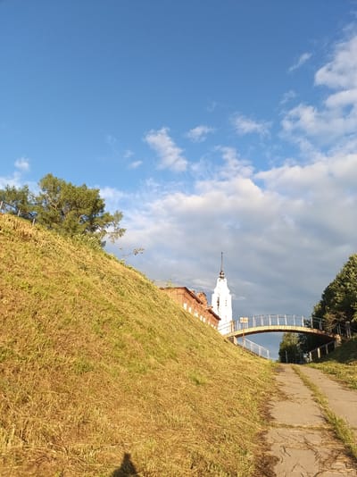 The image depicts a view looking up a grassy slope towards a white church tower with a cross on top, a red brick building, and an arched pedestrian bridge in Buy, Russia. A paved path leads up the hill to the bridge. The sky is blue with scattered clouds. The grass on the slope appears dry and golden, suggesting a warm season. A shadow of a person is visible at the bottom of the slope, indicating a photographer's presence.