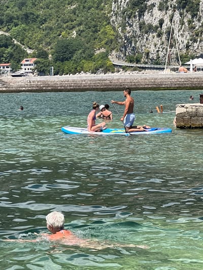 The image shows a sunny day in the Bay of Kotor, near Risan, Montenegro. In the foreground, an elderly person with gray hair is swimming in clear, green water, creating ripples. In the mid-ground, two people are on a paddleboard in the shallow water. A man is kneeling on the paddleboard and holding a paddle, while a woman is sitting on the board. Further in the background, two more people are visible in the water. The shoreline features a stone wall, and beyond that, lush green hills with some buildings, including a white house with a red roof, are visible. A white sailboat mast rises above the hillside. The water is calm and reflects the bright sky and surrounding landscape.