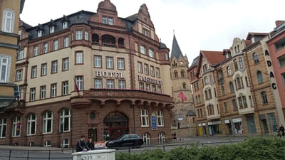 The image depicts the Hotel Kaiserhof in Eisenach, Germany. The hotel is a large, multi-story building with a tan brick facade and brown stone detailing, featuring multiple windows and balconies adorned with flowers. To the right of the hotel, a tall, slender church tower with a clock is visible. Several other buildings, including a pink facade building with "ADAC" signage, line the street. In the foreground, a black SUV is parked in front of the hotel entrance. Two individuals are standing on the sidewalk near the hotel. The scene is set on a street with a traffic light and a grassy median.