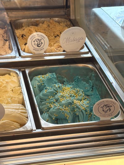 The image shows a close-up view of several metal containers filled with ice cream in a display case. In the foreground, a container holds light blue ice cream swirled and topped with small gold beads. To its left, another container holds pale yellow ice cream. To the left of that, partially visible, is a container with a light brown ice cream.
Two circular labels are visible. One, partially obscured, is on the container with the light brown ice cream, featuring a sheep's head in silhouette and text that includes "OVČIJE MLIJEKO" and "SHEEP MILK". The other label, on the container with the blue ice cream, also features a goat's head in silhouette and text that includes "KOZJE MLIJEKO" and "GOAT MILK". A third label, mostly visible in the upper right, is white and oval-shaped, with "Malaga" written in script, and "MALAGA" and "HOME MADE" in smaller print.
The display case appears to be part of an establishment selling ice cream. The lighting suggests an indoor environment, possibly a shop or gelateria. The background is slightly out of focus but shows more ice cream containers and the reflective surface of the display case. The overall impression is of a chilled display showcasing various flavors of ice cream. The location is not specified in the image itself, and no people are visible.