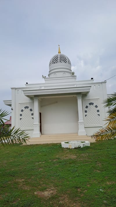A white mosque with a golden dome and a crescent moon sits in the city of Bireuenmeunasahdayah, Indonesia. The mosque has a modern design with clean lines, a large covered entryway supported by columns, and decorative geometric patterns on its facade. Speakers are mounted on the sides of the building, and palm fronds frame the lower portion of the image. A grassy lawn surrounds the mosque, with a small concrete planter in the foreground. The sky is overcast, with a few clouds visible.