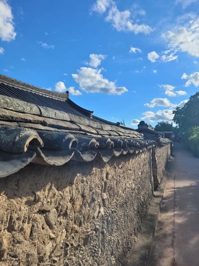 The image shows a stone wall with a tiled roof, likely in Gyeongju-si, South Korea, given the architectural style. The wall is constructed from rough-hewn stones and mortar, and the roof is made of curved, dark tiles. A paved path runs alongside the wall. The sky above is a vibrant blue with scattered white clouds. The scene suggests an outdoor setting, possibly a historical site or a traditional village. There are no people clearly visible in the foreground, but a few figures are present in the background further down the path. The lighting indicates it is daytime, likely late afternoon due to the shadows cast on the path. The overall impression is one of a well-preserved traditional Korean architectural element under a clear sky.
