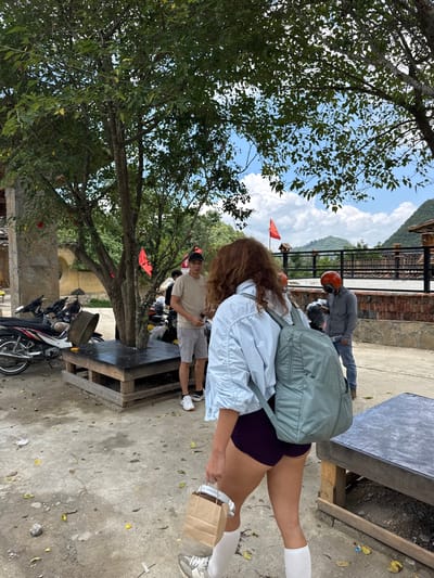 The image, taken in Lũng Cú Commune, Vietnam, captures an outdoor scene. A person with long, reddish-brown hair, dressed in a light blue shirt, dark shorts, and white socks, stands on a concrete surface with a light green backpack. They also hold a brown paper bag in their right hand. Behind this individual, another person in a gray shirt and shorts stands near a motorcycle parking area. An orange helmet is visible on one of the motorcyclists. A third individual wearing a gray jacket and orange helmet is seen near a brick structure. A rustic wooden table with a dark gray top stands in the foreground. A large tree with lush green foliage dominates the left side of the frame. In the background, mountains and a cloudy sky are visible. Red flags are placed in the area. Motorcycles are parked haphazardly. The scene conveys a sense of casual activity within a rural or semi-urban environment.