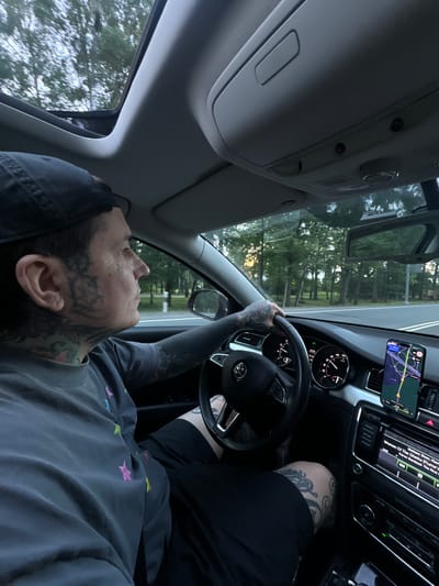 The image shows a person driving a car on a road in Druzhba, Russia. The person is wearing a dark t-shirt and a baseball cap. They have extensive tattoos on their face, neck, and arms. The car's interior is visible, including the steering wheel, dashboard, and a GPS device mounted on the windshield. The road is lined with trees, suggesting a rural or suburban setting. The time of day appears to be late afternoon or early evening, as indicated by the soft lighting. The person is looking forward, focusing on the road ahead.