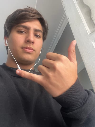 A young man is taking a selfie from a low angle. He is wearing a black sweater and white wired earbuds. His left hand is in the frame, making a "hang loose" gesture. The background includes a white ceiling. The location is unknown and there is no indication it is in Gerli, Argentina.