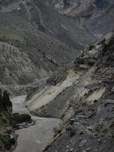 The image depicts a dramatic mountain landscape in the vicinity of Gondhla, India, characterized by a winding river flowing through a deep gorge. The mountainsides are steep and rocky, with sparse vegetation, primarily consisting of scrub and a few scattered trees. A road is visible carved into the mountainside, with vehicles navigating its curves. The river itself is wide and appears to be fast-flowing, with sediment giving its water a muddy appearance. The overall impression is one of rugged, untamed natural beauty, with evidence of human infrastructure in the form of the road. The lighting suggests an overcast or diffused daylight, with muted colors dominating the scene.