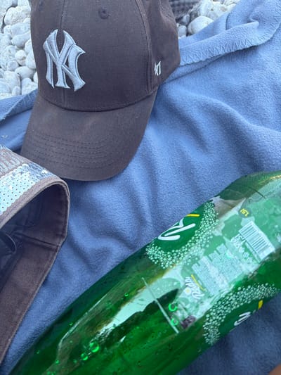 An overhead shot shows a brown New York Yankees baseball cap and a partially visible brown bag with a sequined rim resting on a wrinkled blue blanket. Next to these items is a green plastic bottle of "Pivo" brand beer. The background consists of a collection of small, smooth grey stones, suggestive of a beach or riverbed. The scene is outdoors, possibly in Radovići, Montenegro, given the natural elements.