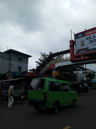 The image captures a bustling street scene in an urban or semi-urban area, likely in Indonesia, under a heavily overcast sky. The lighting is diffused and grey, suggesting either an early morning, late afternoon, or simply a very cloudy day.
In the foreground, a dark asphalt road occupies the bottom portion of the frame. A bright green microbus or minivan, possibly an older model, is the most prominent vehicle, positioned slightly to the right of the center, with its rear facing the viewer. Its windows reflect the cloudy sky. To the left, a person wearing a light-colored hijab, a light brown top, and white pants with white sneakers is walking from left to right along the side of the road. Two motorcycles are parked near a fence behind this person. Further back on the right side of the road, several other people are visible, appearing somewhat blurry due to movement, with one individual wearing a backpack.
The midground features a complex arrangement of buildings and structures. On the left, a multi-story building with a grey and light grey facade and square windows stands. Below it, a smaller, low-roofed structure, possibly a shop or stall, adds a splash of color with its base painted in vibrant blocks of red, blue, yellow, and green, topped with a dark corrugated roof and fronted by a metal fence. This colorful structure appears to form the base of a pedestrian overpass.
The pedestrian overpass itself is a prominent feature, extending from the left towards the right, spanning over the road. It has a yellow railing and black support beams along its stairs and walkway, consistent with the colorful base structure. Behind the overpass and buildings, some green foliage from trees is visible, offering a touch of nature amidst the urban setting.
In the background, on the right side of the frame, a large red and white billboard dominates the upper portion. It features a portrait of a man wearing glasses and a suit, who appears to be a public figure or politician. Visible text on the billboard includes:
- "MARI SU..." (likely part of "MARI SUKSES" or a similar phrase, meaning "Let's succeed").
- A logo with some text below "MARI SU...".
- Large bold text: "KEJU..." and below it "DO...".
- Below the portrait, smaller text names are visible: "HARY TANOESUDIBJO" and "RIDOY SAHRUL".
- Partially obscured large text below the names: "PENGGO..." (possibly "PENGGOLONGAN" or related).
The scene depicts everyday life with pedestrians and vehicles moving under an overcast sky, suggesting a typical day in this bustling area. No strong emotions are discernible, and the overall atmosphere is one of routine activity.