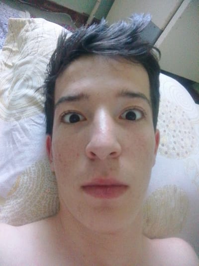 A close-up, selfie-style photograph shows a person's face from the chest up, lying in bed. The person has dark, spiky hair and fair skin with some redness and blemishes. Their eyes are wide open, and their mouth is slightly parted, with visible red lips. The background consists of a pillow with a patterned fabric and a portion of what appears to be a wooden structure above. The lighting is soft and diffused. The image does not contain any elements or context that would allow for a determination of its location in Bukhara, Uzbekistan.