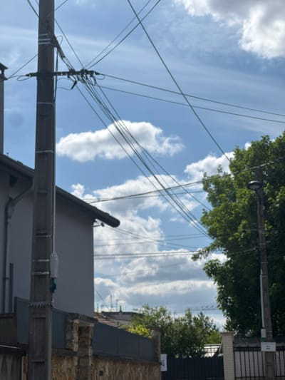The image depicts an urban scene in Champigny-sur-Marne, France. Overhead power lines crisscross a partly cloudy sky. The tops of buildings are visible, constructed of stone, grey, and black materials. Green trees line the upper edges of the scene. A utility pole anchors the power lines.