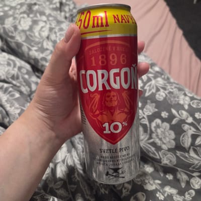 A person is holding a Corgon beer can in a close-up shot. The can is primarily red and yellow, with text indicating "500ml NAVY", "ZALOŽENÉ V ROKU 1896", and "CORGOŇ" in large, bold lettering. Below "CORGOŇ" is an illustration of a muscular man, and further down, "10%" is displayed. The bottom of the can features the text "SVETLÉ PIVO HRDO NESÚCE MENO LEGENDÁRNEHO NITRIANSKEHO KOVAČA JURAJA CORGOŇA" along with an anvil, hammer, and tongs. The background consists of a bed with grey floral patterned bedding and a pink blanket. The image does not provide specific details about a location in the city of Piešťany, Slovakia.