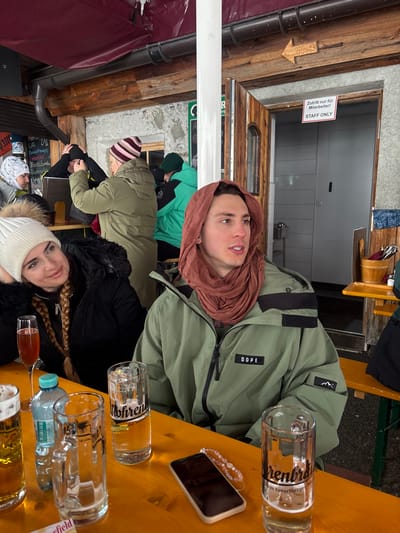 The image captures a semi-outdoor seating area of a establishment in St. Christoph, Austria, featuring rustic wooden and stone construction under a dark red awning. In the foreground, two young adults are seated at a light-brown wooden table. A female on the left, wearing a white knitted beanie and a black winter coat, looks towards the male on the right with a slight smile. The male wears a sage green winter jacket with "DOPE" branding and has a reddish-brown scarf draped over his head and neck. He is looking to the right, off-camera.
On the table are three large beer steins (two partially filled with clear liquid, one labeled "Mohrenbräu das Vorarlberger Bier", and one containing an amber liquid), a flute glass with a reddish-pink beverage, a plastic bottle of "Römerquelle" water, and a smartphone with a light-colored case and a beaded accessory lying face down. In the midground, several other individuals are visible standing and interacting near a counter area, including a person in an olive-green coat and a striped beanie, and another in a bright teal jacket. The background features a textured stone wall, a wooden door with a small window, marked "Zutritt nur für Mitarbeiter! STAFF ONLY," and a small wooden arrow sign pointing right above the door. Overhead, wooden beams and a rain gutter system are visible.