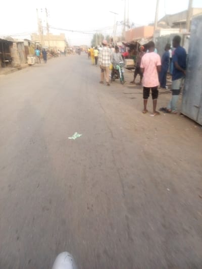 A straight asphalt road runs through a town. On both sides, various people are walking and standing. One man is on a small motorcycle. Some buildings and shops line the edges of the road, with utility poles visible in the distance. Litter lies on the road. The overall lighting suggests either early morning or late afternoon.