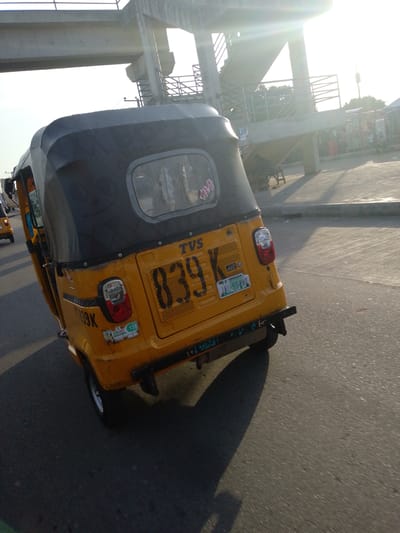 The image captures a yellow auto-rickshaw driving on a paved road, possibly in Gbaza, Nigeria. The auto-rickshaw, a common mode of transport in many parts of the world, is tilted slightly to the left, indicating movement. The vehicle is adorned with a black tarp-like covering over its passenger compartment. Prominently displayed on the back of the auto-rickshaw are the license plate numbers "039 K" and "JAL-554DT". The background reveals an overpass structure, possibly part of a bridge or elevated road, with a sun glare obscuring parts of the sky. Vegetation and some buildings are visible in the distance, suggesting an urban or semi-urban environment. The overall impression is a candid shot of daily life in a bustling Nigerian city, with the auto-rickshaw being the primary focus.