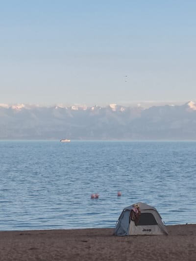 The image depicts a serene lakeside scene in Bayet, Kyrgyzstan. In the foreground, a sandy beach is visible, with a light gray Jeep-branded tent set up on the sand. Clothing and a hat are draped over the entrance of the tent. Two pink buoys float on the calm, blue water of the lake. In the middle ground, a white boat is visible on the water. The background features a hazy, majestic mountain range with snow-capped peaks, under a clear blue sky.