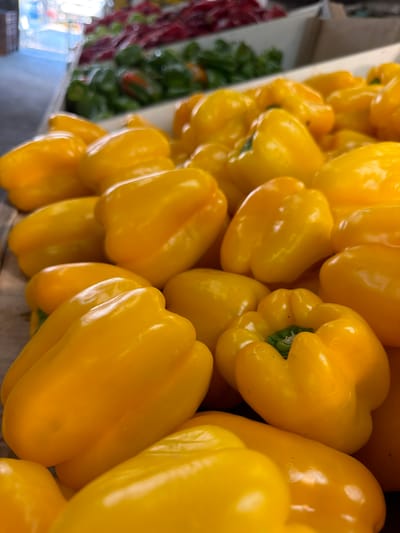 A close-up, slightly angled shot of a display of bright yellow bell peppers, likely at a market or produce stand. The peppers are tightly packed together, creating a dense arrangement. Some peppers are fully visible, while others are partially obscured. In the blurred background on the left, glimpses of other produce, including red items and leafy greens, can be seen. The location appears to be a market, possibly in Kadima-Zoran, Israel, based on contextual information.