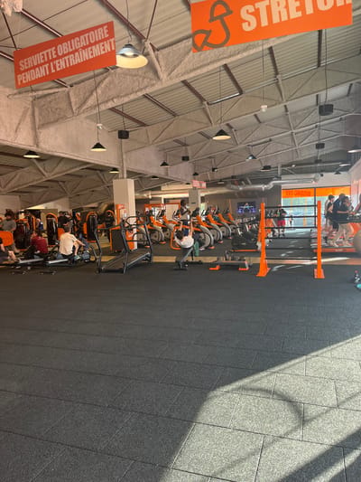 The image depicts the interior of a Basic-Fit gym in Illkirch-Graffenstaden, France. Rows of orange exercise bikes are occupied by individuals engaged in workouts. Overhead signage in French is visible, including one for "STRETCH" and another stating "Serviette Obligatoire Pendant L'entrainement" (Towel Required During Training). The gym features a high ceiling with visible support beams and ventilation. The floor is a dark, textured material. Sunlight streams in from windows at the far end of the gym.
