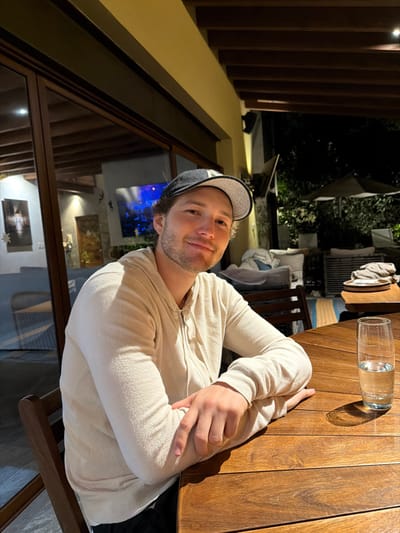A male individual is seated at a dark brown wooden slatted table on a covered outdoor patio at night in Valle de Bravo, Mexico. The subject, a light-skinned male with short brown hair and stubble, wears a dark baseball cap with a white emblem and a cream-colored, full-zip hooded sweatshirt. He faces the camera directly with a slight smile, his forearms resting on the table. To his right on the table is a clear glass containing water.
The environment includes a wooden chair matching the table. To the left, behind glass sliding doors, an interior space with light-colored walls, wooden ceiling beams, and artwork is visible, including a framed print of a tree-lined path, another piece of art, and a large screen displaying blue and white abstract visuals. A small star ornament hangs near the interior artwork. To the right, beyond the seated individual, an outdoor area is illuminated by ambient light against the darkness of night. This area contains cushioned outdoor seating, a small table with stacked items, and two large, folded umbrellas partially obscured by dark foliage. The patio ceiling consists of wooden beams with recessed lighting fixtures.