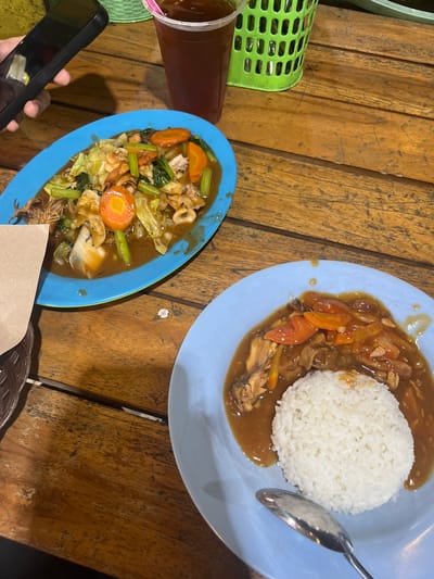 Two plates of food are shown on a wooden table. The plate in the foreground contains a mound of white rice and a dish of meat in a brown sauce with sliced carrots and tomatoes. A silver spoon rests on the plate. The plate in the background contains a stir-fry dish with various vegetables, including cabbage, carrots, and celery, and pieces of seafood, possibly squid and shrimp. A hand holding a black smartphone is visible in the upper left corner. A plastic cup filled with a dark liquid and a pink straw is positioned behind the plate in the background. A green plastic basket with a woven pattern is visible to the right of the cup. The table surface is made of wooden planks with visible grain and wear. The setting appears to be a casual eatery, likely in Boja, Indonesia, given the context of the location.