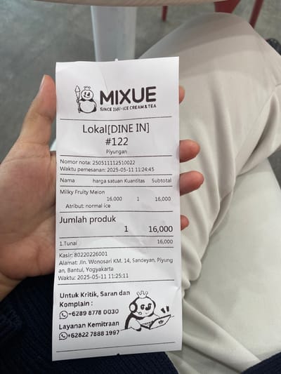 The image is a receipt from Mixue, an ice cream and tea shop. The receipt is held in the right hand of a person.  The location of the store is Lokal [DINE IN] #122, Piyungan, Indonesia.  The receipt number is 25051112510022 and the time of purchase was 2025-05-11 11:24:45. The customer purchased one Milky Fruity Melon with normal ice for 16,000 Indonesian Rupiah. The total cost of the order was 16,000 Indonesian Rupiah. The cashier's number is 80220226001 and the store address is Jln. Wonosari KM. 14, Sandeyan, Piyungan, Bantul, Yogyakarta. The time of the transaction was 2025-05-11 11:25:11. The receipt also includes contact information for complaints and feedback.  The background is blurry and features a white surface.  The receipt is printed on white paper with black ink.