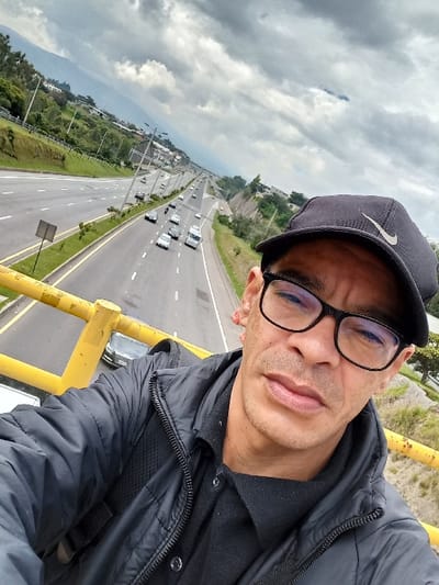 An adult man wearing glasses and a black baseball cap poses for a selfie. He is wearing a black jacket over a collared shirt. Behind him is a highway with multiple lanes of traffic, various cars are visible. There is some green vegetation, buildings, and mountains in the distance. The sky is cloudy. The man is leaning against a yellow painted barrier.