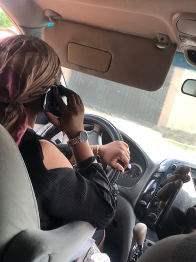 A partial interior view of a vehicle, from a rear passenger perspective, shows an adult driver in Egan, Nigeria. The driver, with dark skin, wears a beige and reddish-pink patterned headwrap and a black, ribbed, long-sleeved top with an exposed shoulder. Her left hand, wearing a rose gold-toned wristwatch and a thin gold-colored bracelet, grips the black-covered steering wheel. Her right hand holds a dark-colored mobile phone, possibly a feature phone, to her right ear, indicating an active conversation. Dark nail polish is present on both hands. The vehicle's interior features a beige dashboard and a sun visor in the upright position. The automatic gear selector is in the "D" (Drive) position, with a light brown knob. Through the front windshield, a light-colored building or fence with horizontal lines is visible, along with some green foliage. A patterned item, possibly a bag, is partially visible on the center console or front passenger seat.