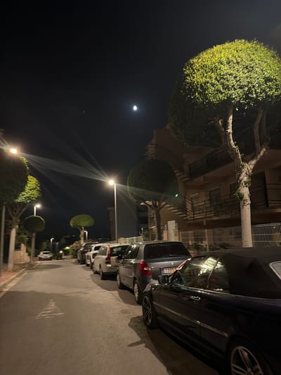 The image depicts a street scene at night in Cambrils Badia, Spain. The street is lined with parked cars and illuminated by streetlights. The moon is visible in the dark sky. Several trees with rounded canopies are also present along the street, and a building with balconies and an external staircase is visible on the right side of the image. The overall impression is a quiet residential street at night.