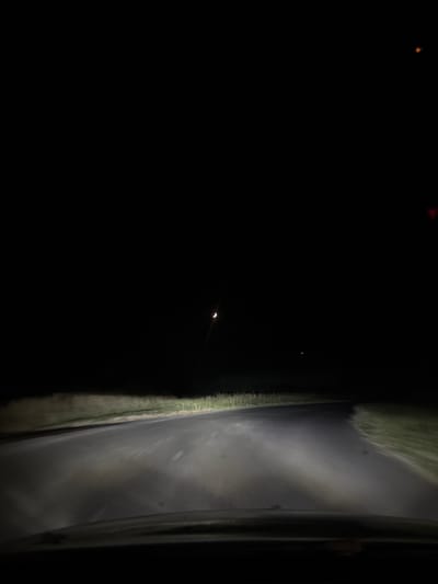 The image depicts a nighttime view from a vehicle driving on a rural road. The road is paved and curves to the right. Headlights illuminate the road surface and the grassy verges on either side. The sky is dark, with a crescent moon visible and a distant light source, possibly a streetlight or another vehicle's lights. There are no people or other discernible subjects in the image. The scene appears to be located in an unpopulated area, consistent with the outskirts of a town or village. The presence of a paved road suggests it is a thoroughfare, possibly leading into or out of Kableshkovo, Bulgaria. The overall atmosphere is dark and solitary.