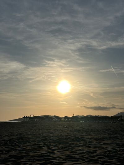 A long beach in Donji Štoj, Montenegro is captured at sunset. The sun is shining brightly behind thin clouds, creating a golden hue across the sky. The dark, sandy beach stretches out to the right side of the image, exhibiting patterns of wind-blown ripples. Along the horizon, there are structures such as beach umbrellas, sunbeds, and buildings. In the sky, a faint contrail from an aircraft is visible.