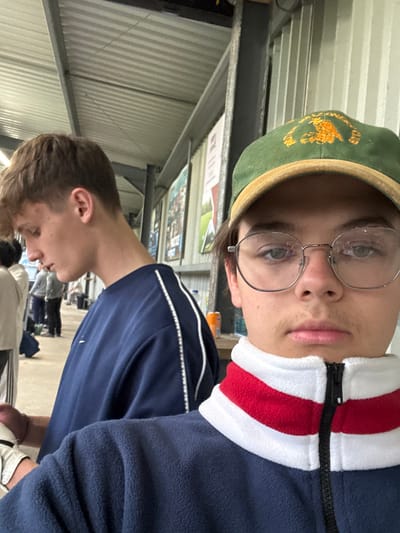 The image shows two young men standing in an indoor sports facility, likely a cricket training center, located in South Ribble, United Kingdom at the coordinates 53.771, -2.57. The man on the left is wearing a navy blue sweatshirt with white trim and is looking down. The man on the right is wearing a green and yellow baseball cap, clear framed glasses, and a navy blue fleece jacket with red and white trim. He is looking directly at the camera. There are other people visible in the background, including a couple of people who appear to be wearing cricket gear. The facility appears to be a typical indoor cricket training facility, with a high ceiling and walls that are decorated with posters.